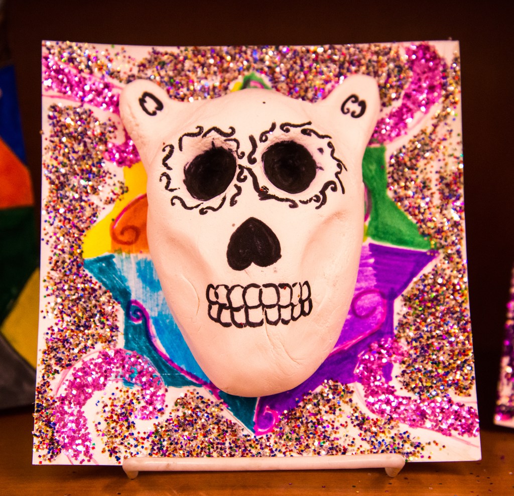 An example of a 4th grade student's clay skull, designed to imitate a Día de los Muertos (Day of the Dead) sugar skull, with a colorful and glittery patterned background