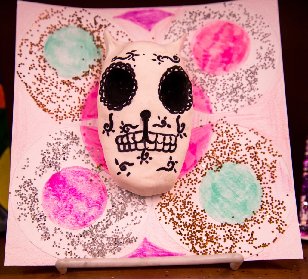An example of a 4th grade student's clay skull, designed to imitate a Día de los Muertos (Day of the Dead) sugar skull, with a colorful and glittery patterned background