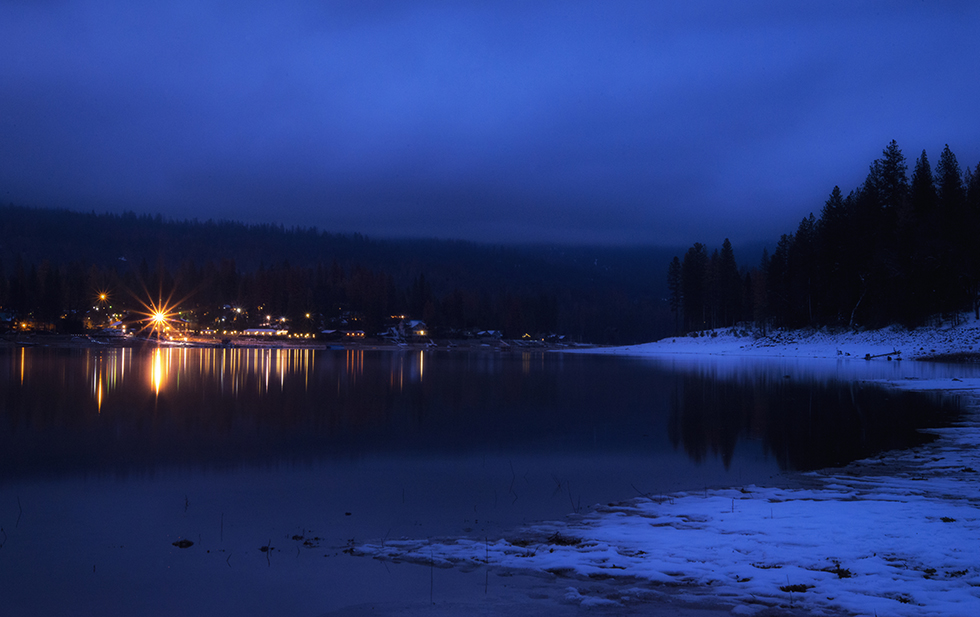 Snowy winter scene, with the lights from homes reflecting on he water's edge, in Bass Lake, California, by Anita Sagastegui