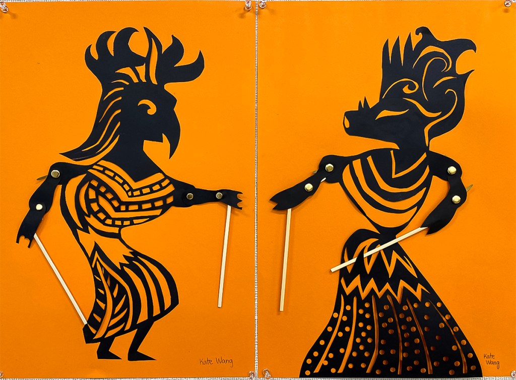 An 8th grade student's paired Wayang Kulit puppets. The puppets are made of black paper and set against a bright orange background to look like shadow puppets. They are intricately perforated to create elaborate patterns within the costume of each puppet, as well as to express each puppet's features. Their arms are joined with brass fasteners at the elbow and shoulder, manipulated at the hands by sticks.