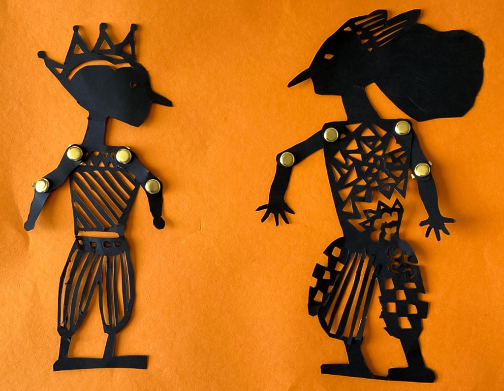 An 8th grade student's paired Wayang Kulit puppets. The puppets are made of black paper and set against a bright orange background to look like shadow puppets. They are intricately perforated to create elaborate patterns within the costume of each puppet, as well as to express each puppet's features. Their arms are joined with brass fasteners at the elbow and shoulder, manipulated at the hands by sticks