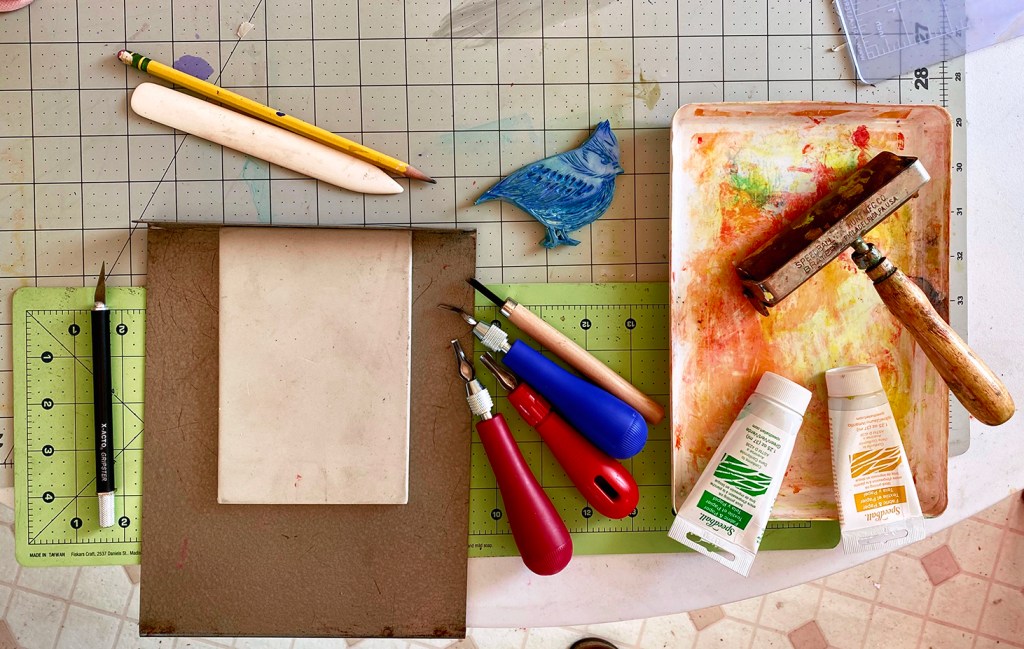 A snapshot of some necessary supplies for linoleum block printmaking: linoleum block, bone folder, carving tools, Xacto knife, brayer, paint, paint tray, self-healing craft mat, non-slip carving tray.