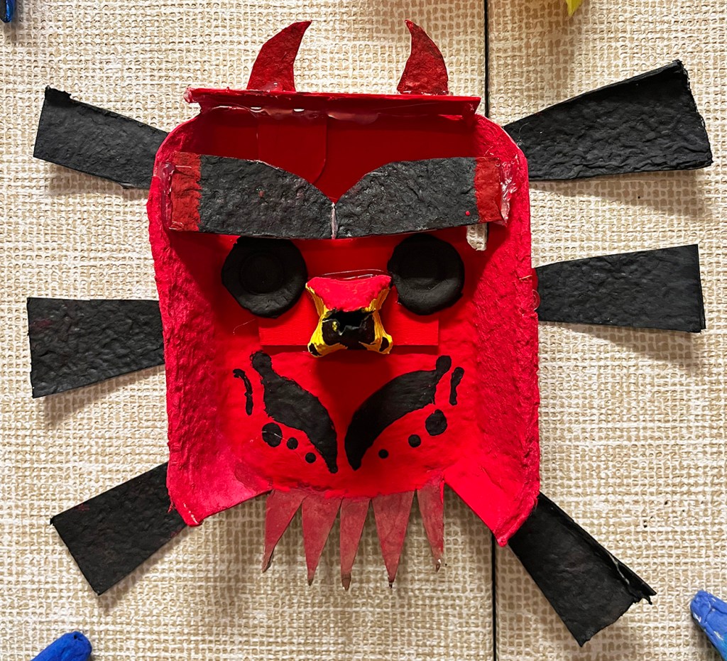 A 6th grade student's masks made of egg cartons, inspired by masks of the pacific Northwest tribes. This one has some head decorations and is painted in mostly red and black.