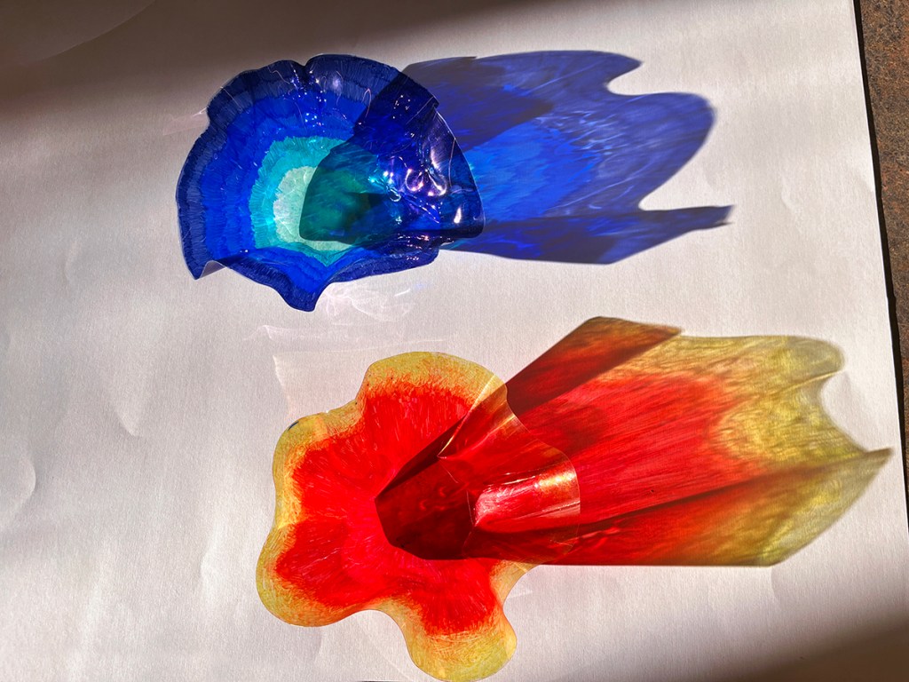 Melted plastic bowls shaped in the style of Dale Chihuly's glass bowls, by 8th grade students. Two bowls, one in vibrant shades of blue, the other bowl in vibrant yellow, orange and red