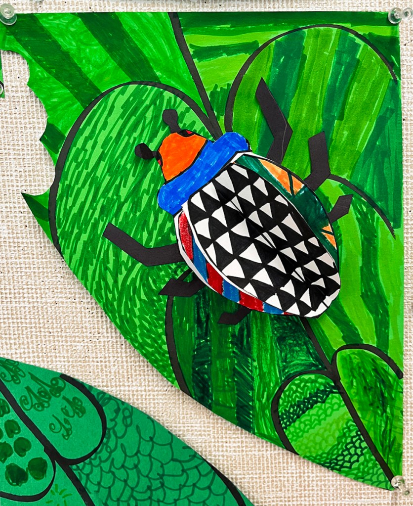 Three-dimensional bug, with vivid colors and diverse patterns on the wings, crawling across a green leaf that itself is covered in varied patterns, created by first grade students