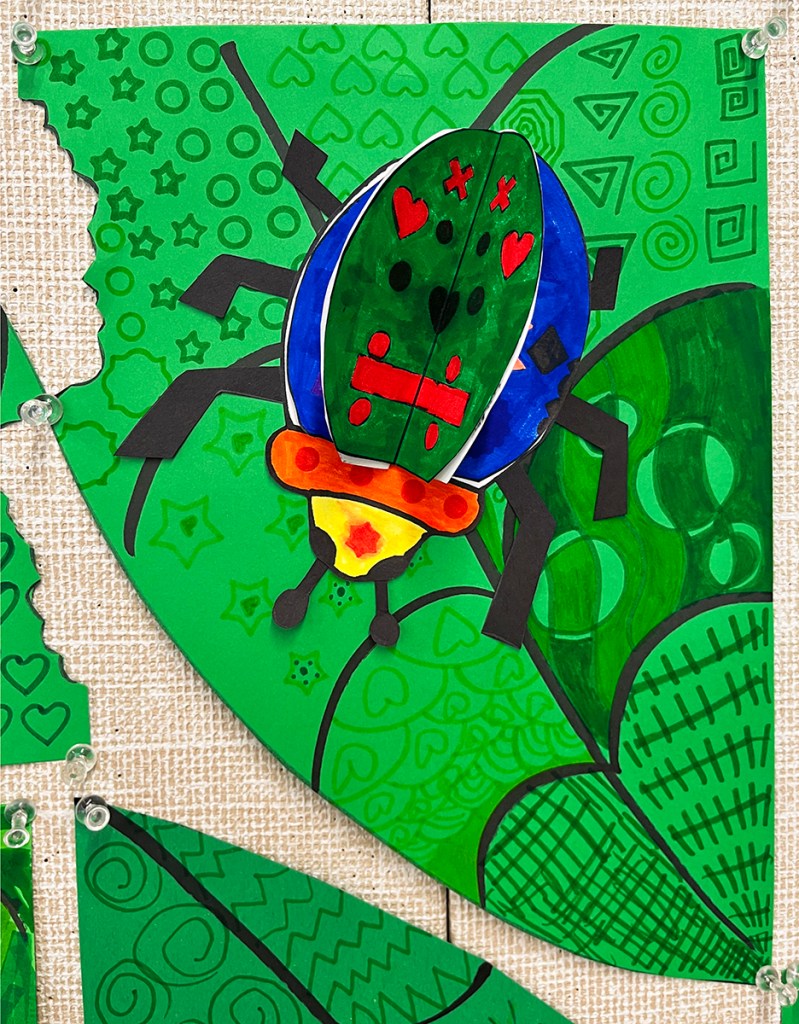 Three-dimensional bug, with vivid colors and diverse patterns on the wings, crawling across a green leaf that itself is covered in varied patterns, created by first grade students