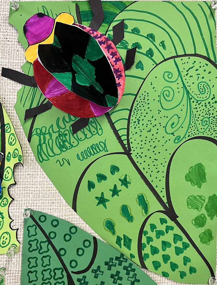Three-dimensional bug, with vivid colors and diverse patterns on the wings, crawling across a green leaf that itself is covered in varied patterns, created by first grade students