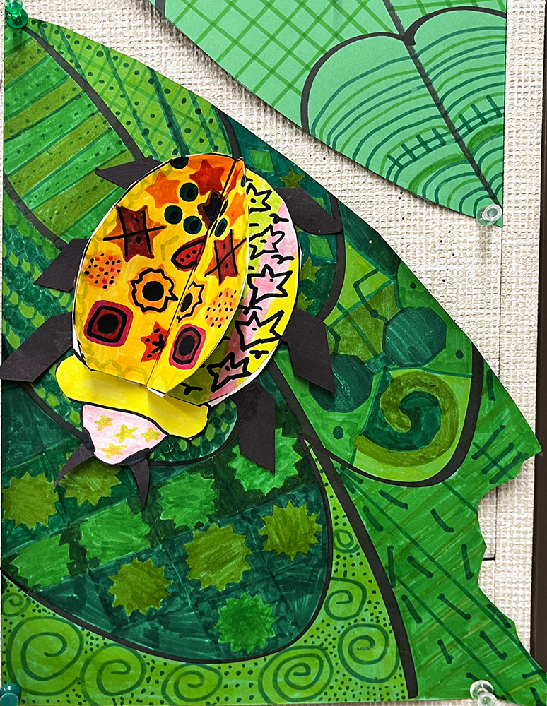 Three-dimensional bug, with vivid colors and diverse patterns on the wings, crawling across a green leaf that itself is covered in varied patterns, created by first grade students